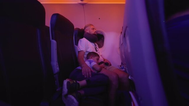 Father wearing neck pillow and young son sleep peacefully in airplane seats during overnight flight. Purple and orange ambient cabin lighting creates cozy atmosphere for rest.