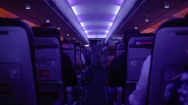 Interior shot looking down narrow airplane aisle with passengers seated on both sides, atmospheric purple and pink LED mood lighting glowing from curved ceiling panels, overhead compartments visible.
