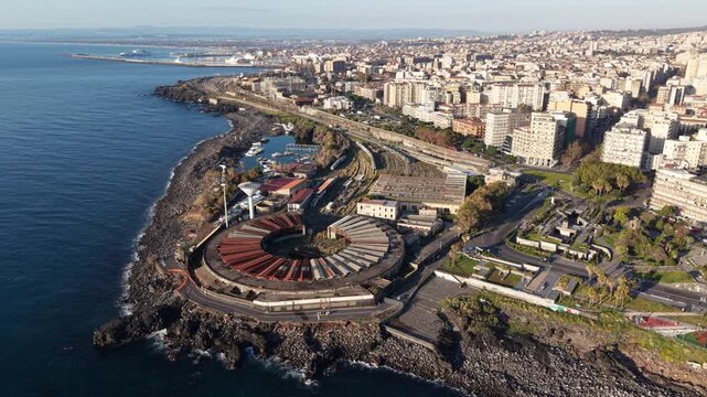 Aerial view of the circular railway depot on the volcanic rocky coast with the urban skyline and blue sea in Catania, Sicily, Italy.