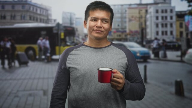 Man holding a red enamel mug and smiling while standing on a busy city street with buses, cars and pedestrians in a casual travel setting; calm contentment.