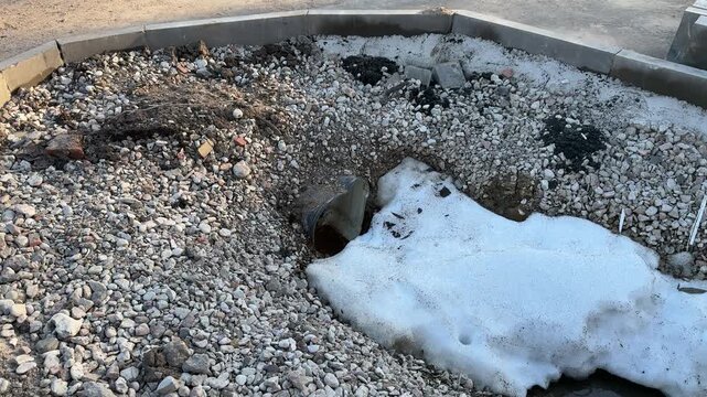 Snow on crushed stone around drain, concrete rim, asphalt edge, small sewer pipe opening with melting runoff and silt, pale winter