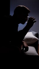 Fototapeta premium Silhouette of a man awake at midnight in a dark bedroom, sitting alone and drinking a glass of water while wrestling with insomnia, anxiety and quiet loneliness