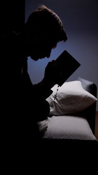 Silhouette of person in quiet reflection holding a bible with rosary draped across it, evoking solitary prayer, faith, humility and peaceful spiritual devotion in dim light