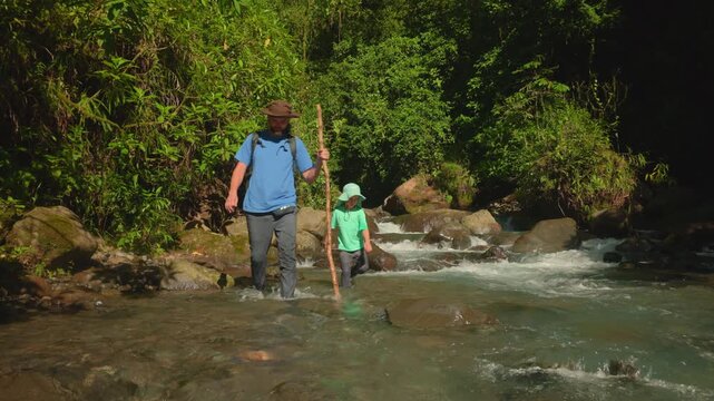 Son with father hiking through wild nature as they wade across a flowing mountain river surrounded by lush tropical forest. Dad uses walking stick while child follows through clear water.
