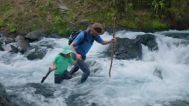 Son with father hiking through wild nature as they carefully wade across a fast-flowing mountain river. Dad holds childs hand while both use walking sticks for balance in rushing white water rapids.