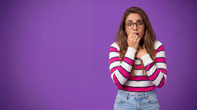 A young woman with glasses and a striped sweater bites her nails, expressing fear or anxiety against a purple background