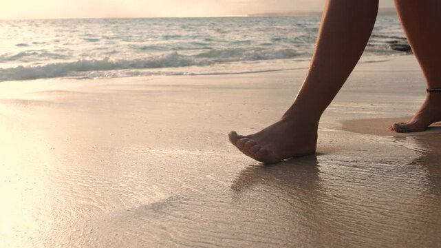 SLOW MOTION, LENS FLARE, CLOSE UP: Golden light glows across wet sand as bare feet of a young woman step along the shore. Tropical beach walk and a peaceful sunset atmosphere in the Mentawai Islands.