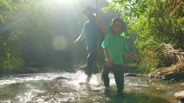 Son with father hiking through wild nature as they wade through a shallow forest stream. Sunlight creates lens flare through dense green foliage while the child leads the way through flowing water.