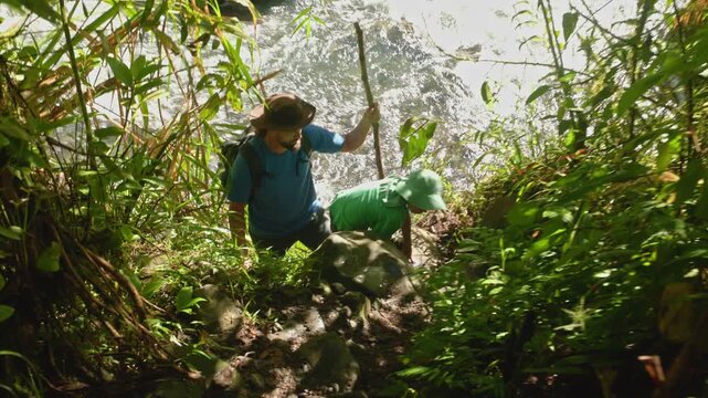 Son with father hiking wild nature as they climb rocky terrain near flowing river. Parent and child explore dense jungle vegetation together using walking stick for support on steep trail.