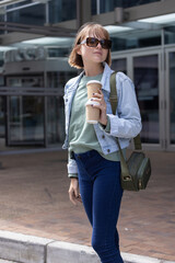 Fototapeta premium Female standing on brick curb at glass entrance wearing denim jacket holding stacked takeaway cups