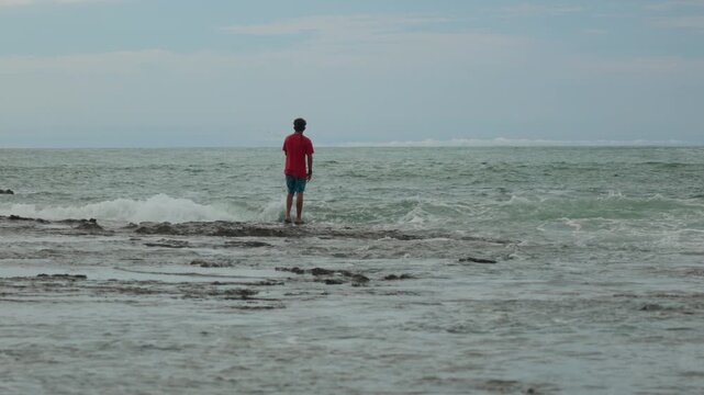 A young man in red shirt and blue shorts stands on a rocky outcrop in the ocean while fishing. Waves crash around him as he casts his line into the turquoise sea under overcast skies.