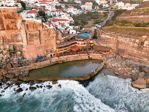 Aerial view of Praia das Azenhas do Mar with white houses on a cliffside overlooking a natural swimming pool with people and crashing waves in Colares, Lisbon, Portugal.