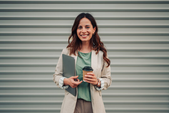 Young businesswoman smiling, holding a laptop and take away coffee cup, standing against a modern urban background
