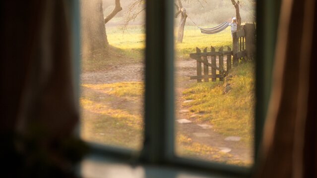 Blurry window frame creating a sense of coziness and privacy, looking out at a person relaxing in a hammock during a sunny summer day in a rustic garden