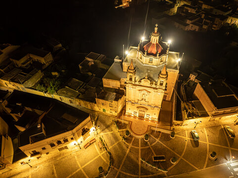 Aerial view of the illuminated Knisja Parrokkjali ta' Munxar church and surrounding village square at night Il-Munxar, Regjun Ghawdex, Malta.