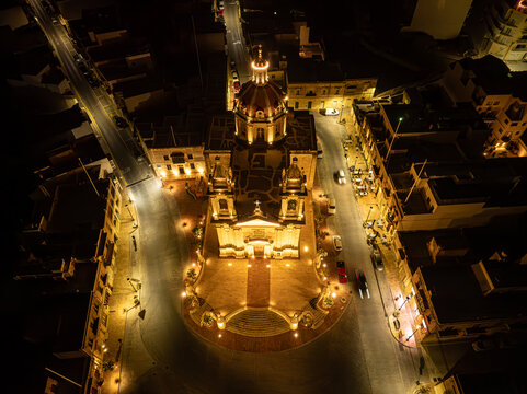 Aerial view of the illuminated Sannat Parish Church with its glowing dome and bell towers overlooking the quiet night streets of Sannat, Gozo Region, Malta.
