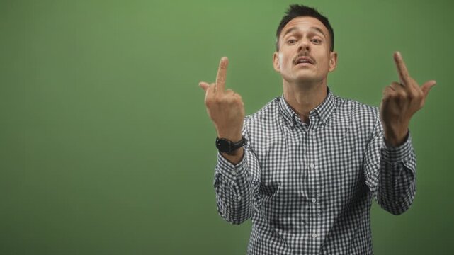 Hispanic man flipping middle finger with both hands in green studio; defiance irreverence contempt attitude.