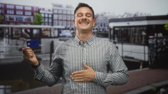 Young hispanic man holds creditcard in raised left hand and places right hand on stomach in street; financial confidence joy.