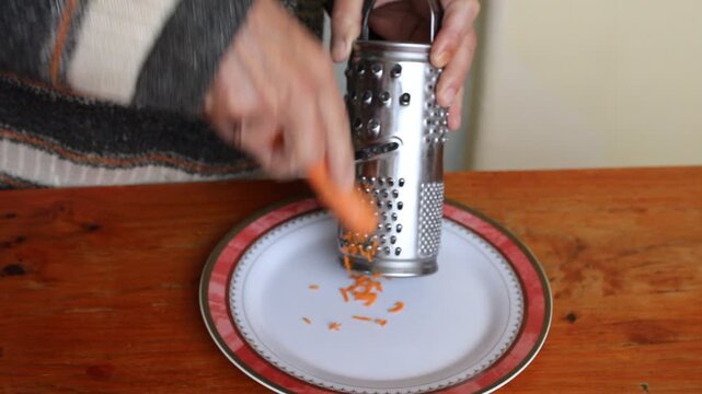 El hombre est&aacute; rallando una zanahoria mientras la sostiene con la mano. Utiliza un rallador de acero inoxidable para triturarla, y los restos caen directamente sobre un plato colocado en una mesa.