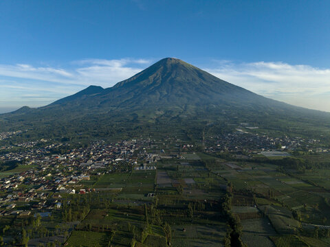 Aerial view of Mount Sindoro rising above the rural landscape, agricultural fields, and residential buildings under a clear blue sky Kecamatan Kertek, Jawa Tengah, Indonesia.