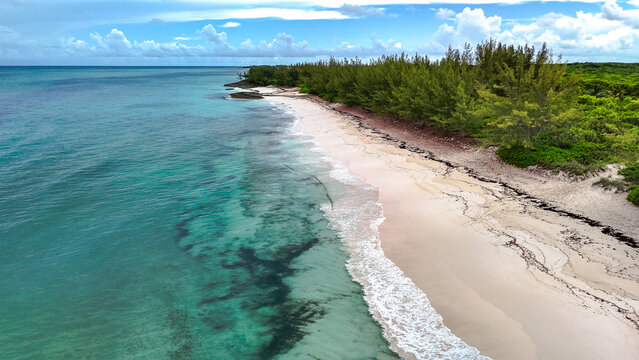 Aerial view of Preacher's cave beach with turquoise waters, white sand, and lush green pine trees under a cloudy blue sky The Bluff Settlement, North Eleuthera, The Bahamas.