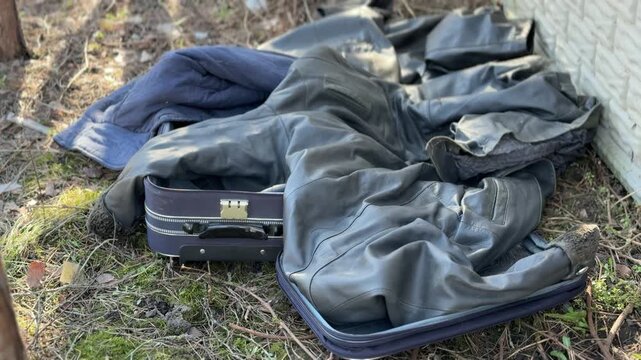 Discarded jacket, trousers, suitcase by fence spring park scene with blue hat on damp grass, sunlit shadows, tree trunk at edge