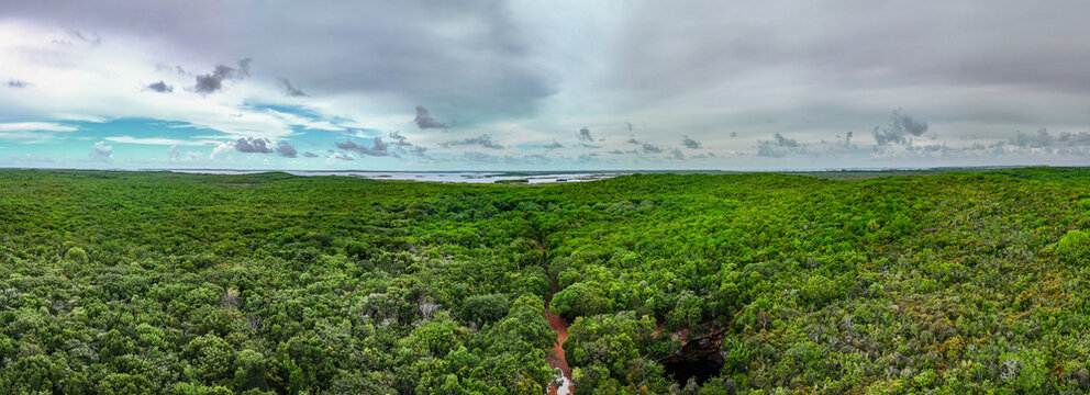 Aerial view of the Sapphire Blue Hole surrounded by dense tropical forest and a dirt road under a cloudy sky in The Bluff Settlement, North Eleuthera, The Bahamas.