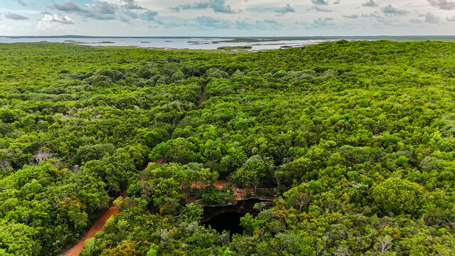 Aerial view of the Sapphire Blue Hole surrounded by dense tropical forest and a dirt path under a cloudy sky in The Bluff Settlement, North Eleuthera, The Bahamas.