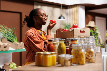 Obraz premium Black woman shops at local market, supporting local producers and choosing eco friendly products like fresh fruits and vegetables. African American lady near the glass checking out the tomatoes.