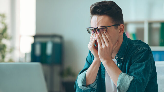 Worried Young Caucasian Man Wearing Glasses Rubbing Eyes With Tired Expression At Office Desk, Struggling With Workload In Modern Office Setting. Business Concept