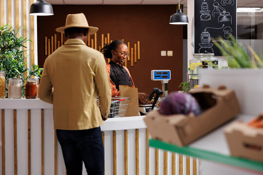 A friendly female cashier with a big smile on her face and a cool-looking male customer wearing a hat. They seem to be having a pleasant interaction at the checkout counter in a grocery store.
