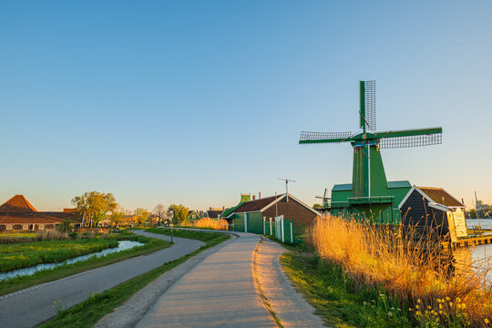 Scenic dutch landscape with traditional green windmill in zaanse schans at sunset, winding countryside road along water canal, historic wooden houses and famous holland tourism landmark