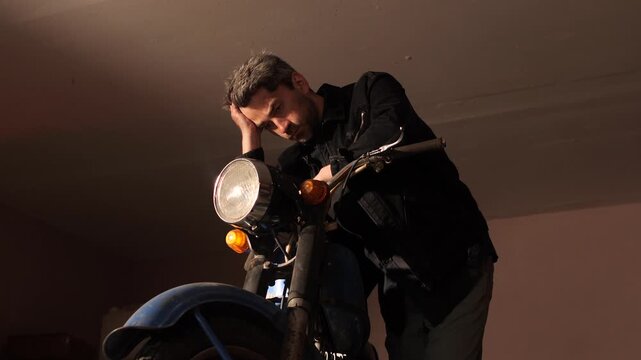 Man with a thoughtful expression leaning on a vintage motorcycle, contemplating deep thoughts in a dimly lit garage, reflecting on challenges or future plans