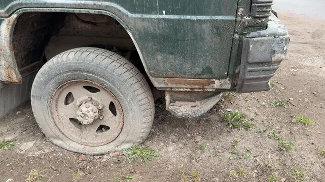 Abandoned dark green minibus punctured tire, rusted undercarriage and chipped paint, spare tires stacked beneath body, moss