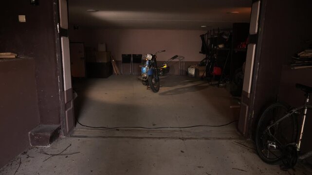 Dark garage interior storing an old blue motorcycle, black bicycle and scattered boxes on a dusty concrete floor, conveying neglected storage, workshop and utility space