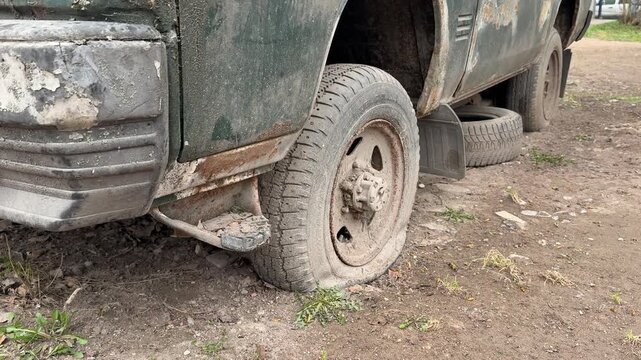 Abandoned dark green minibus punctured tire, rusted undercarriage and chipped paint, spare tires stacked beneath body, moss