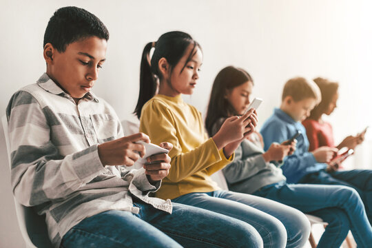 Focused Unrecognizable Group Of Five Kids Including Boys And Girls Sitting Together Using Smartphones In Indoor Setting. Engaged With Mobile Devices, Displaying Modern Technology Addiction Concept