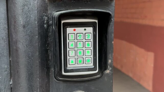 Analog intercom keypad on iron gate, weathered black frame, green backlit numeric buttons, round scanning key below keypad, brick