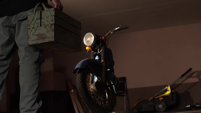 Person walking with a vintage metal toolbox, preparing to work on a classic blue motorcycle or moped in a dimly lit garage workshop, symbolizing a hobby, maintenance, or diy project