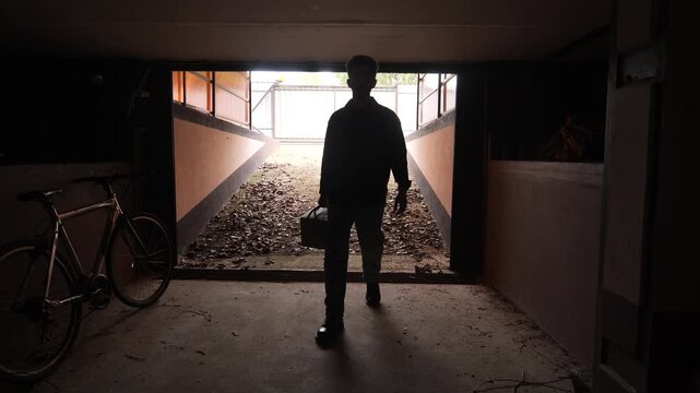 Man silhouette standing inside a dark garage holding a toolbox, preparing to exit towards the bright light, creating a sense of mystery and transition
