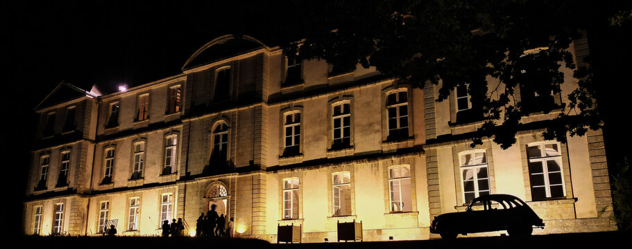 Loyat, France - 01 September 2023: Aerial view of the illuminated Chateau de Loyat at night with a vintage car parked in front and people gathered near the entrance.
