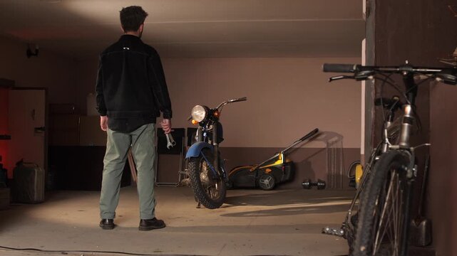 Man holding a wrench in a dim garage, inspecting a classic motorcycle beside a lawnmower, evoking diy repair, hobby workshop, maintenance and thoughtful evening work