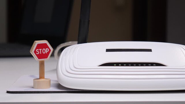 Person's hand interacting with a miniature stop sign placed on a white desk next to a wireless internet router, symbolizing concepts of security, network control, and pausing online activity