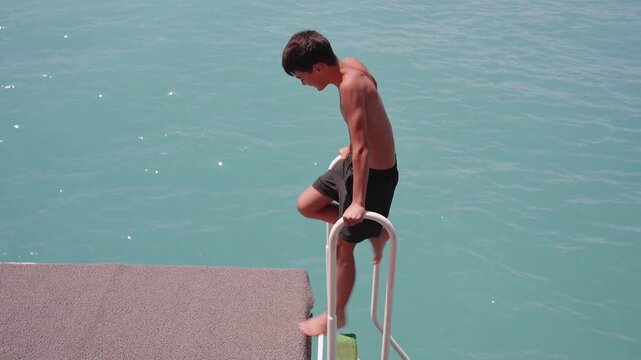 Teenage boy stepping down from pier ladder into clear turquoise sea during sunny beach vacation. Summer travel, swimming, freedom and seaside adventure concept. Happy kid playing on pier