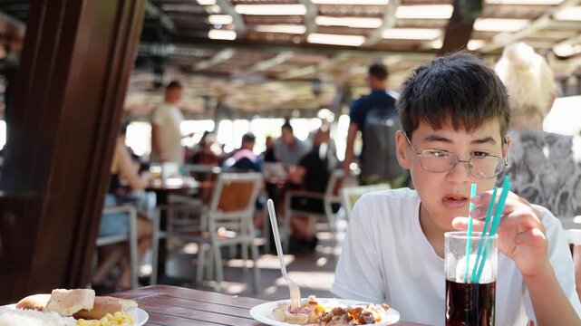Teen boy drinking cold soda through straw while eating lunch at outdoor resort restaurant. Summer family vacation, travel dining and leisure lifestyle concept