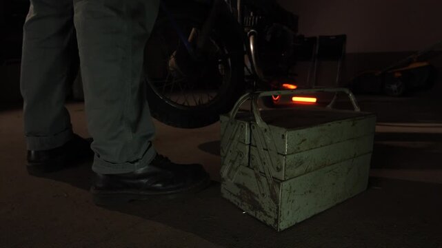 Person standing near a motorcycle with a vintage metal toolbox on the ground, representing repair, maintenance, and skilled labor in a dim environment