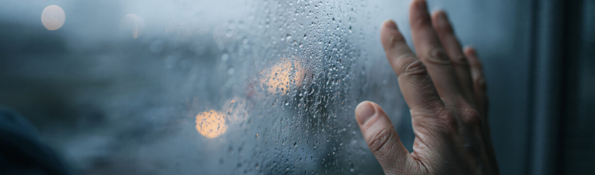 Rainy Day Window View: A person's hand rests on a fogged-up windowpane, with raindrops distorting the view of blurry lights outside, evoking a sense of solitude and introspection.