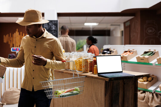 While African American clients browse the store a wireless computer with a blank mockup template is placed on wooden counter. side view shot of a laptop showing an isolated white screen for adverts.