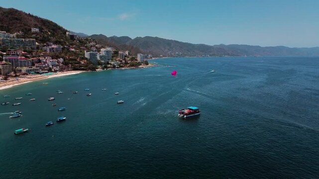 Aerial 4K drone view of a boat towing a parasail over turquoise waters at Los Muertos Beach, Puerto Vallarta, with a ferry, boats and coastal buildings visible in the background