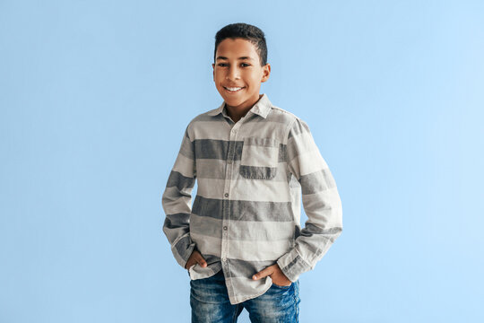Smiling Young Hispanic Boy, Approximately 10-12 Years Old, Standing With Hands In Pockets Against Plain Blue Background, Wearing Gray Striped Shirt In Studio. Child Portrait Photography, Family